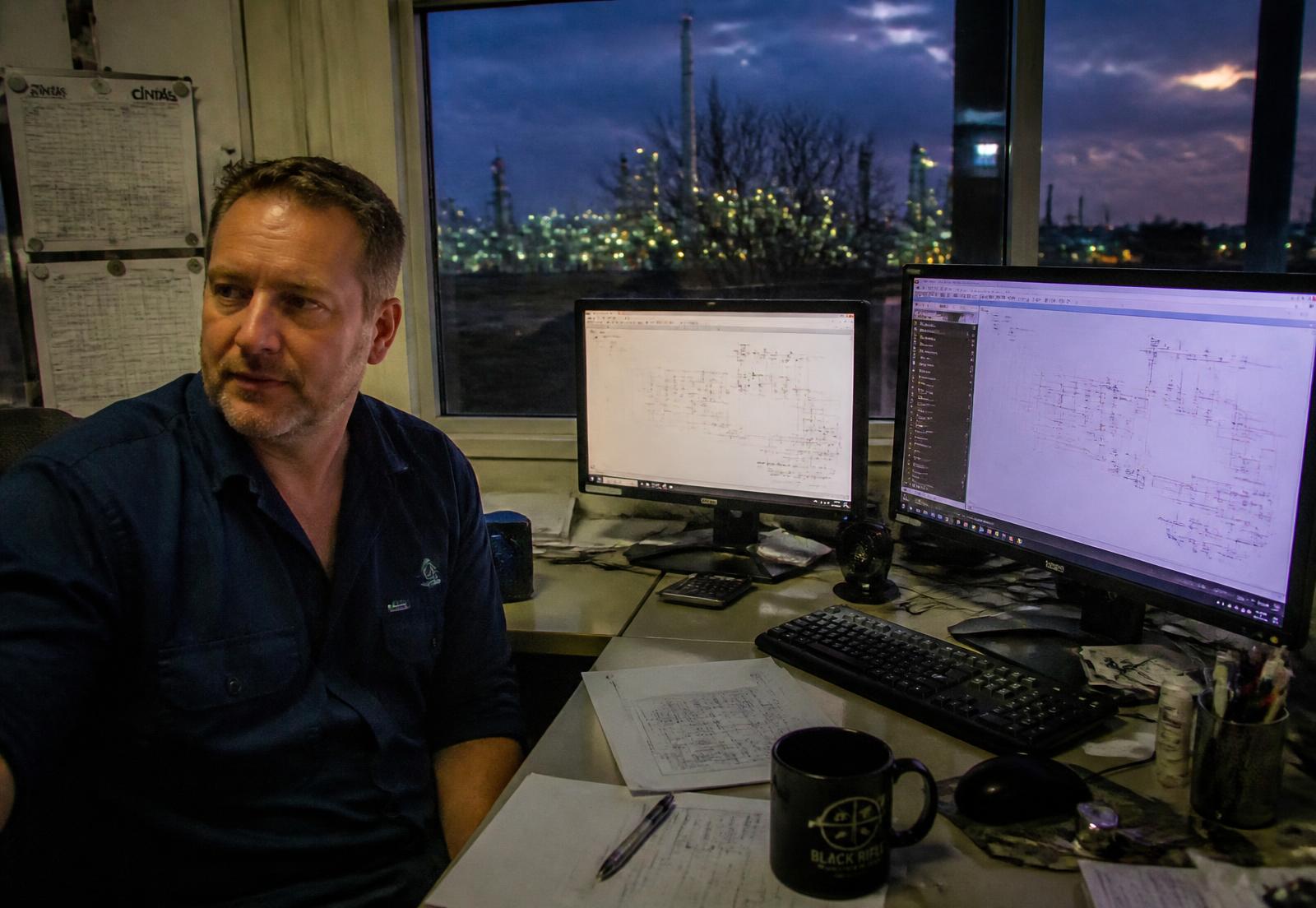 Engineer at a dual-monitor workstation reviewing electrical schematics with an illuminated refinery beyond the window
